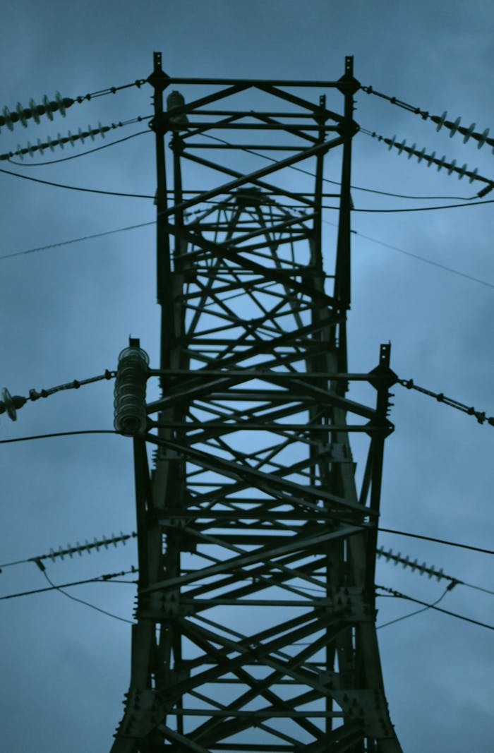 A close-up shot of a high voltage powerline tower silhouetted against a gray cloudy sky.